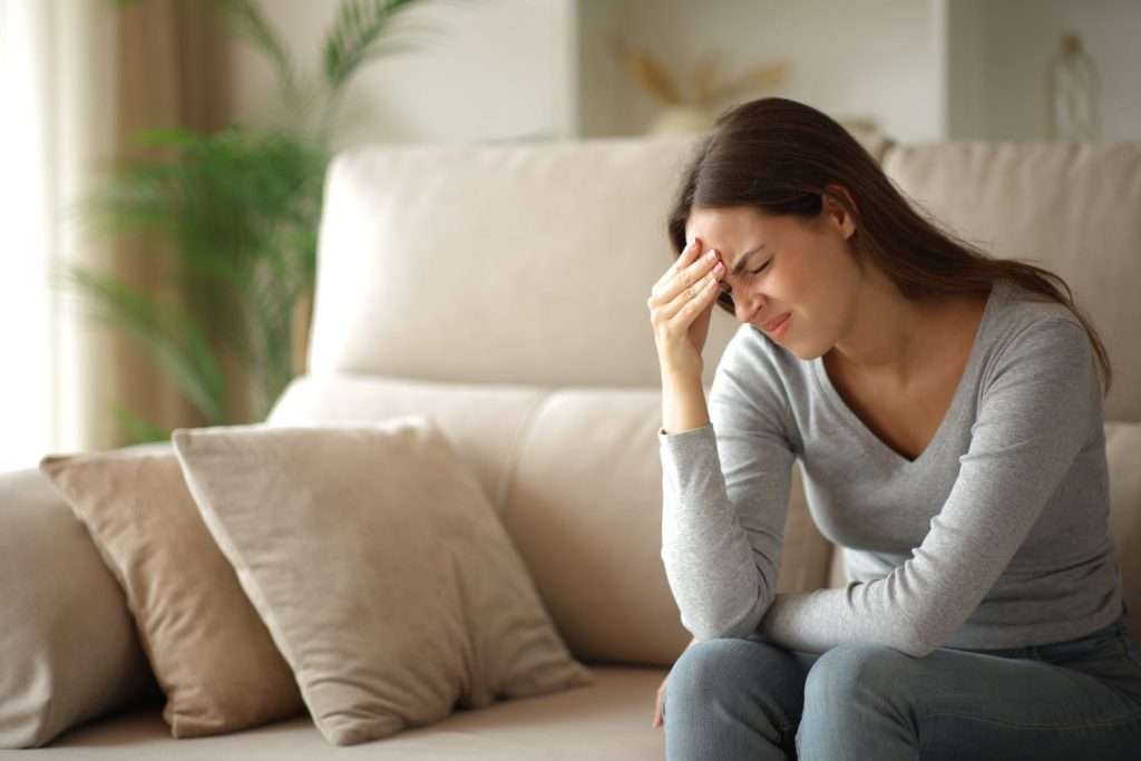 Woman experiencing a severe migraine headache with forehead pain and light sensitivity while sitting on a couch.
